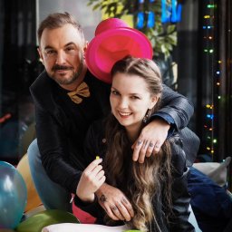 Wedding Assistance Kinga Rek - Uśmiechnięta para siedzi otoczona balonami, mężczyzna w cekinowej marynarce i złotej muszce obejmuje kobietę z różowym kapeluszem na głowie, w tle dekoracje i światełka.