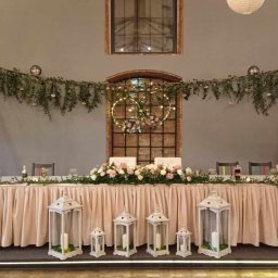 Wedding Assistance Kinga Rek - Uroczysta dekoracja stołu weselnego z białymi lampionami, kwiatami i girlandami z zieleni, wiszące papierowe kule i okrągłe dekoracje świetlne w tle, industrialne wnętrze z ceglanymi ścianami.