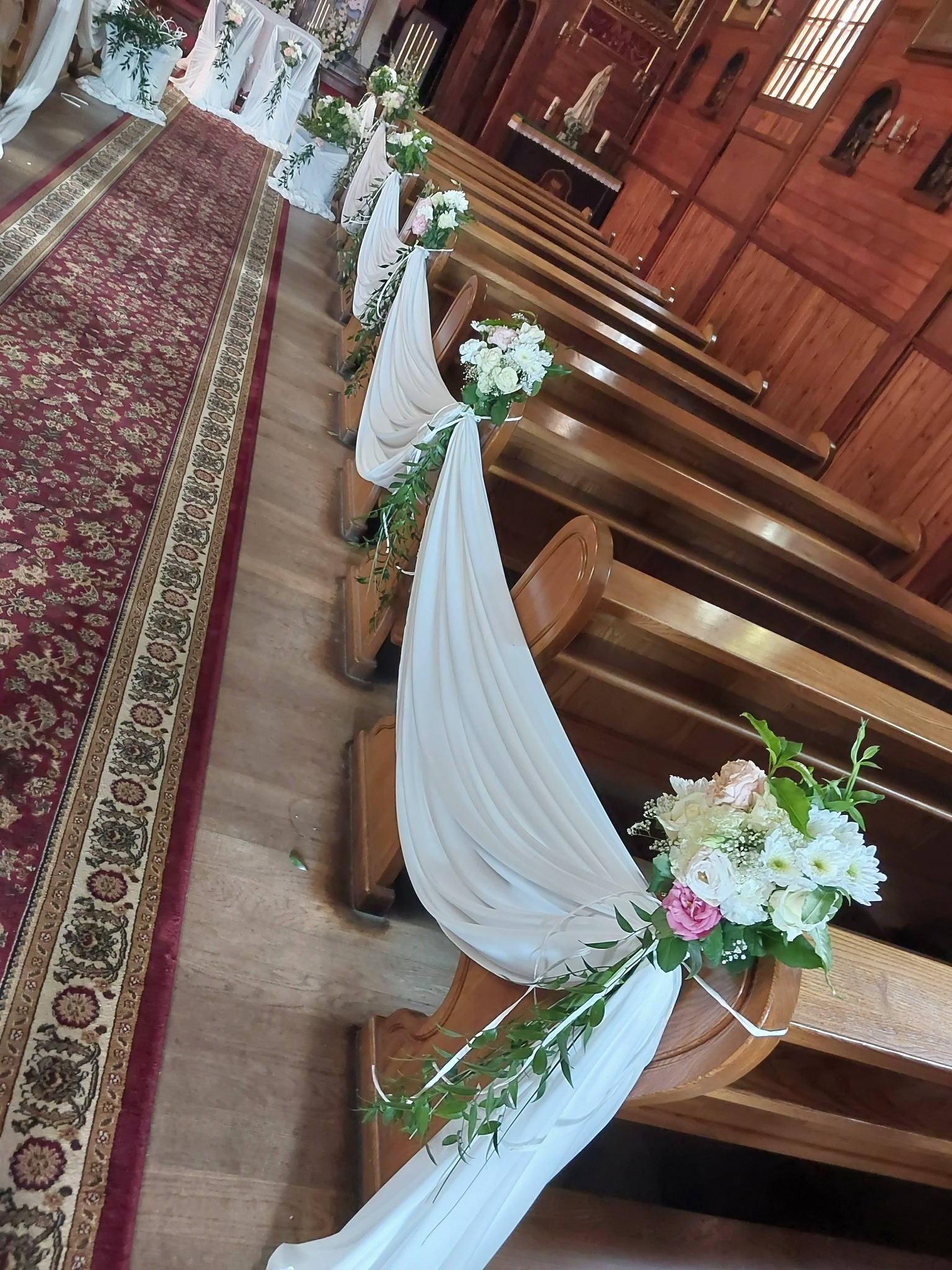 Dekoracja kościoła na ślub: białe draperie i bukiety z białych i różowych kwiatów zdobią drewniane ławki wzdłuż czerwonego dywanu.