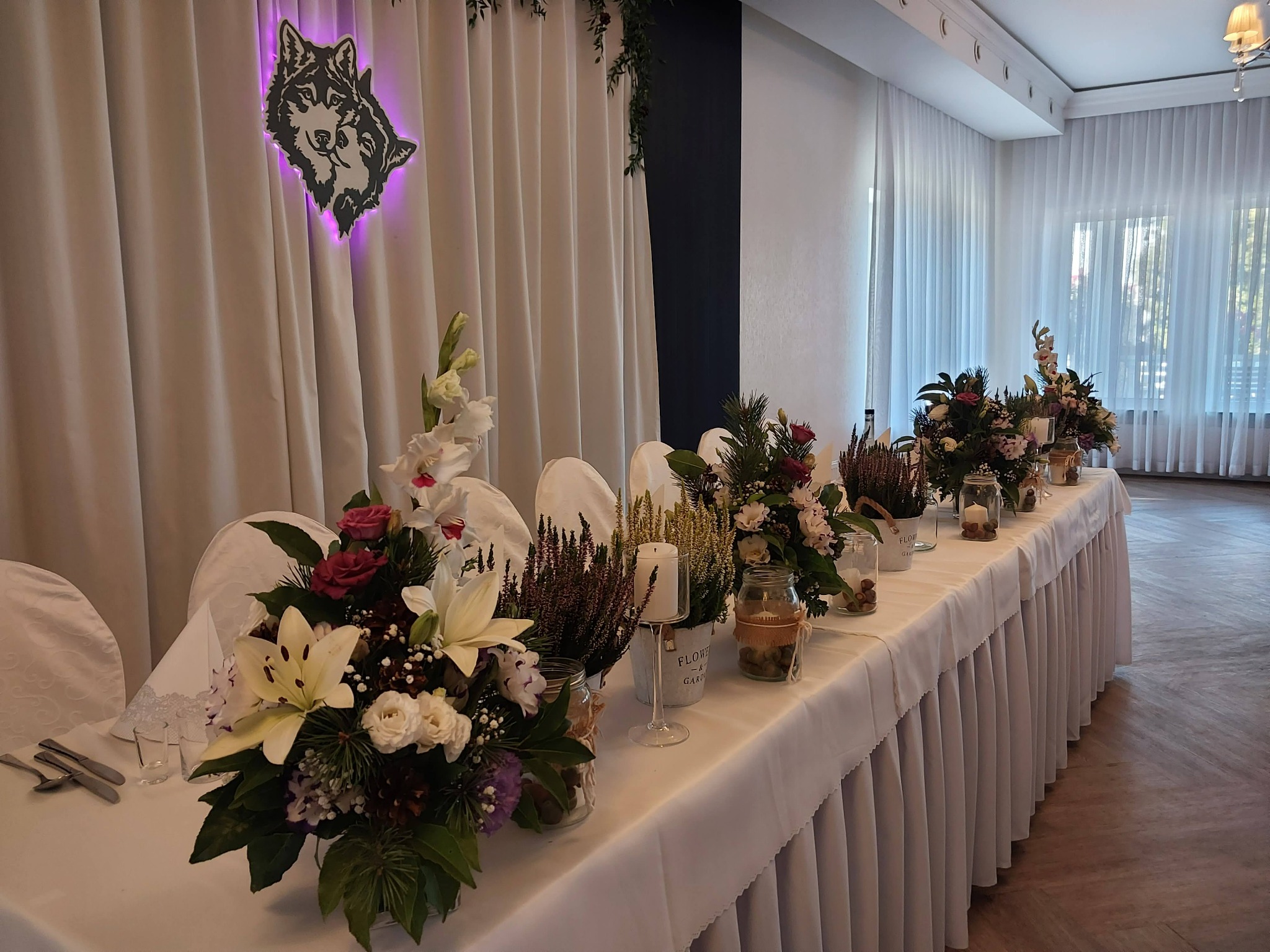 Udekorowany stół weselny z kompozycjami kwiatowymi w wazonach i świecznikach, w tle białe zasłony z podświetlaną grafiką wilków.