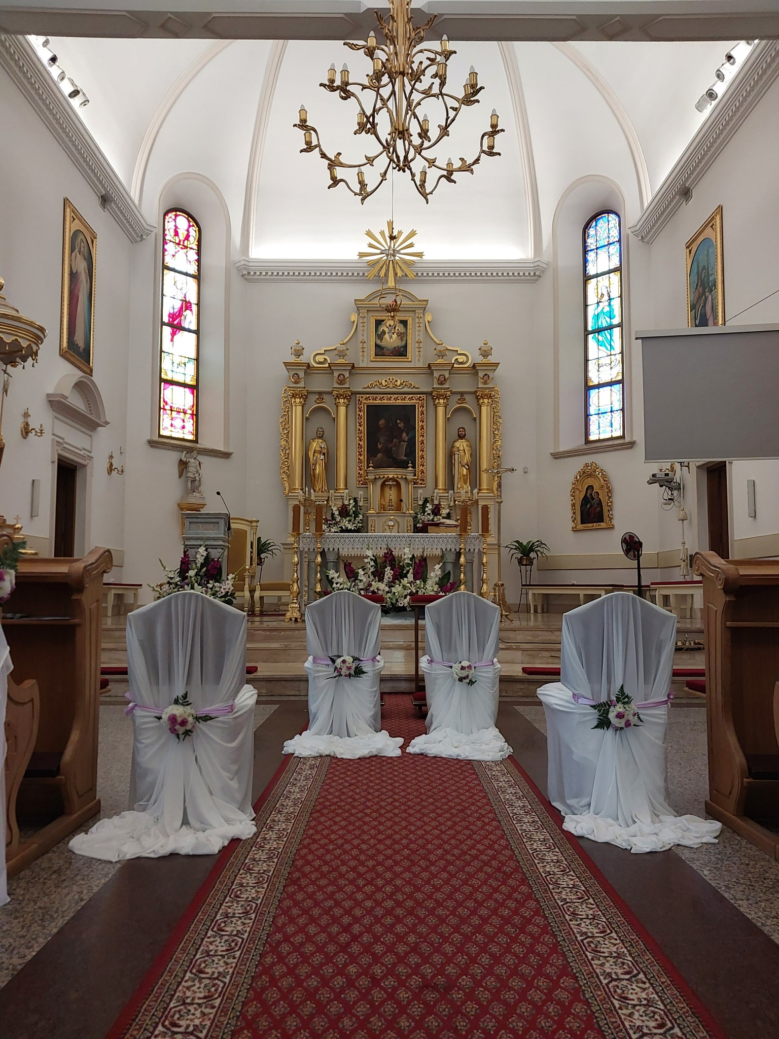 Dekoracja krzeseł w kościele na uroczystość ślubną: białe pokrowce z kwiatami i fioletową wstążką, czerwony dywan z ornamentem, złoty ołtarz w tle, witraże w oknach.