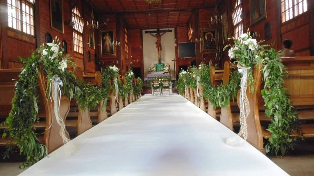 Wnętrze drewnianego kościoła udekorowane zielonymi girlandami i białymi kwiatami na ławkach, biały dywan prowadzi do ołtarza z krzyżem w tle. Ozdobne wstążki zwisają z dekoracji.