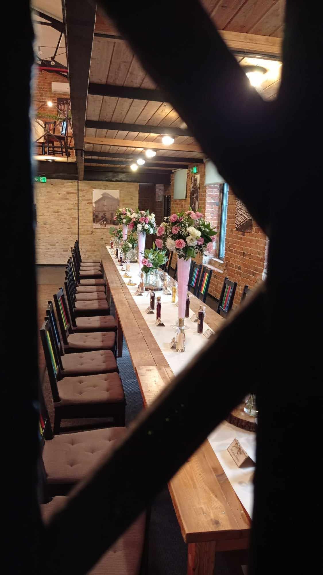 Ustawiony długi stół z drewnianym blatem i białym obrusem, dekorowany kompozycjami kwiatowymi w różowych wazonach i szklanych naczyniach, butelkami z ciemną zawartością i winietkami z nazwiskami...