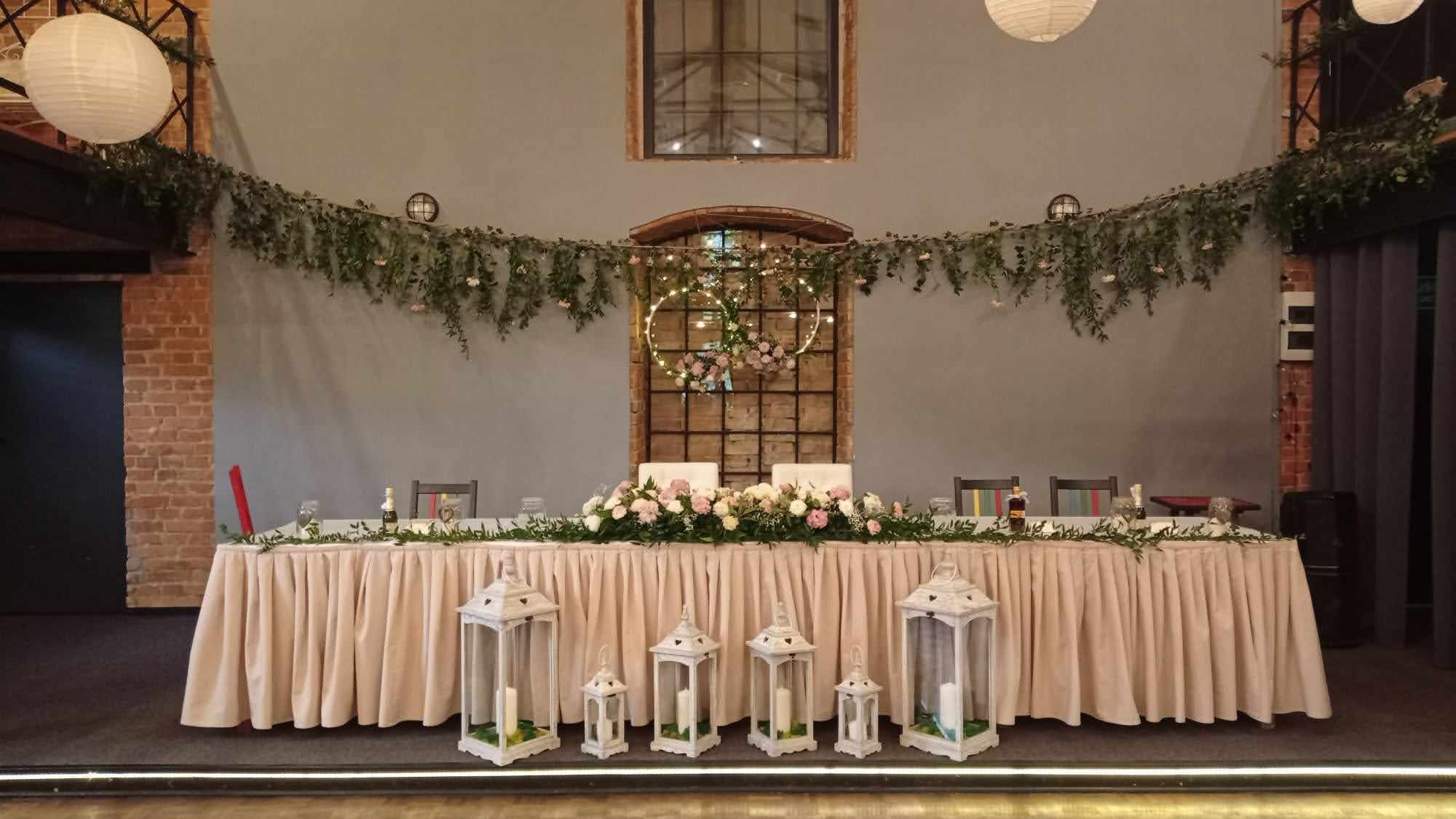 Uroczysta dekoracja stołu weselnego z białymi lampionami, kwiatami i girlandami z zieleni, wiszące papierowe kule i okrągłe dekoracje świetlne w tle, industrialne wnętrze z ceglanymi ścianami.