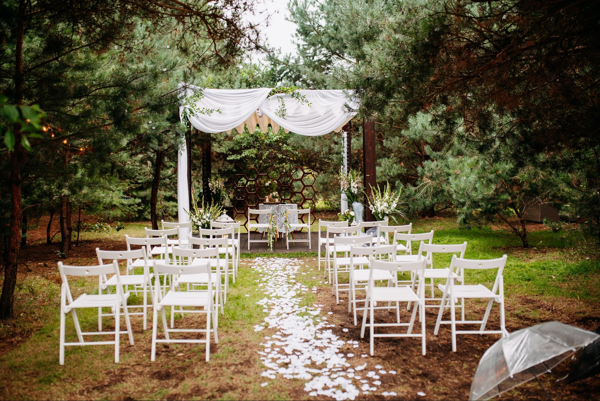 Dekoracja plenerowa ślubu z białymi krzesłami, płatkami kwiatów na ścieżce i drewnianą konstrukcją z białymi draperiami i zielonymi girlandami, otoczona drzewami.