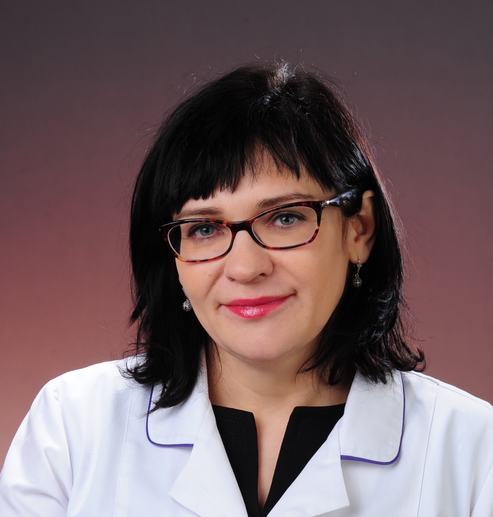 Portret kobiety w okularach i białym fartuchu medycznym z fioletowym obszyciem, na brązowym tle.