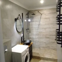 Nowoczesna łazienka z pralką, kabiną prysznicową typu walk-in, umywalką nablatową w kolorze czarnym i okrągłym lustrem z podświetleniem LED.