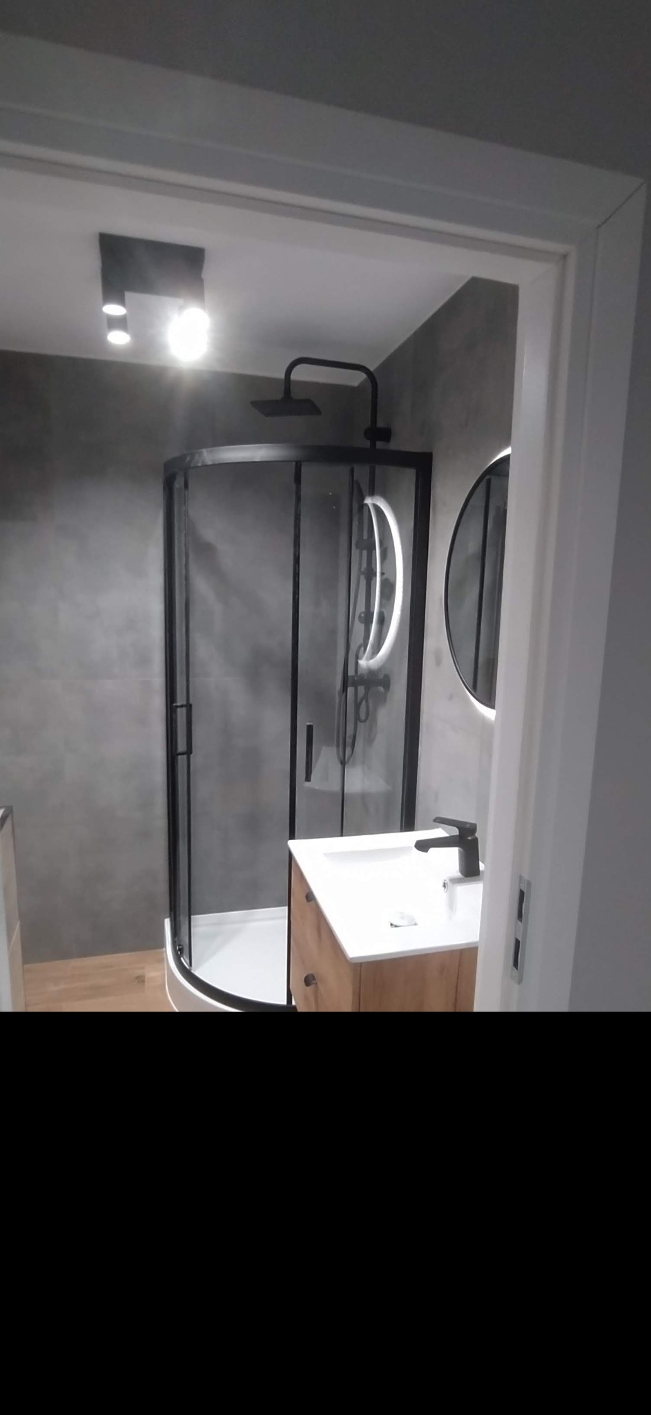 Nowoczesna łazienka z kabiną prysznicową typu walk-in z czarnymi ramami, okrągłym lustrem z podświetleniem LED i drewnianą szafką pod umywalką z czarną baterią, na tle szarej ściany.