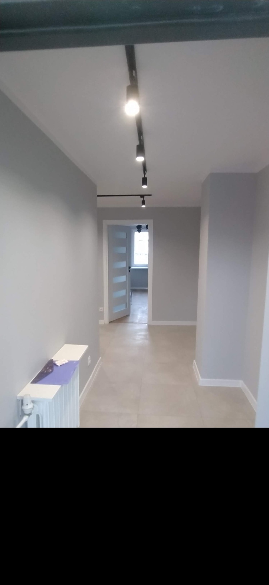 Wnętrze korytarza po remoncie z szarymi ścianami, jasną podłogą, grzejnikiem z białym blatem i nowoczesnym oświetleniem szynowym.