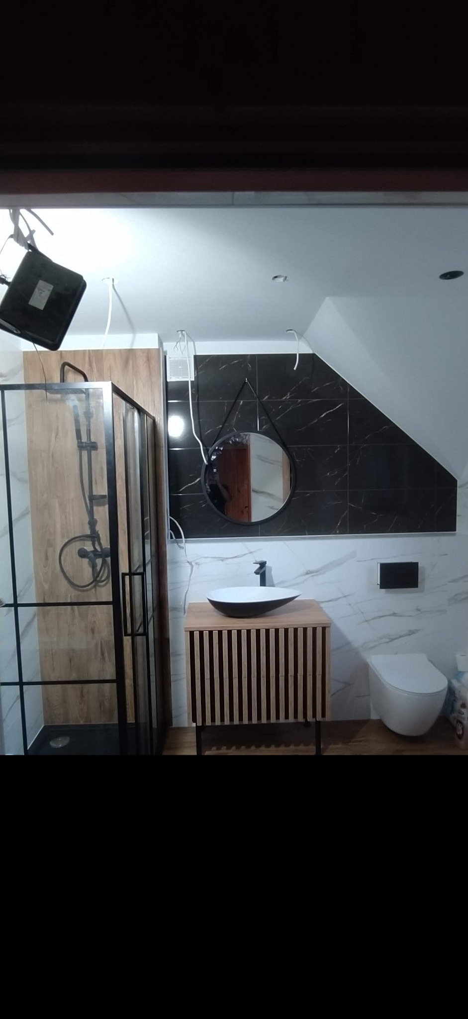Nowoczesna łazienka w trakcie wykańczania: kabina prysznicowa z czarną ramą, umywalka nablatowa na drewnianej szafce z lamelami, okrągłe lustro i toaleta wkomponowane w biało-czarne marmurowe płytki.