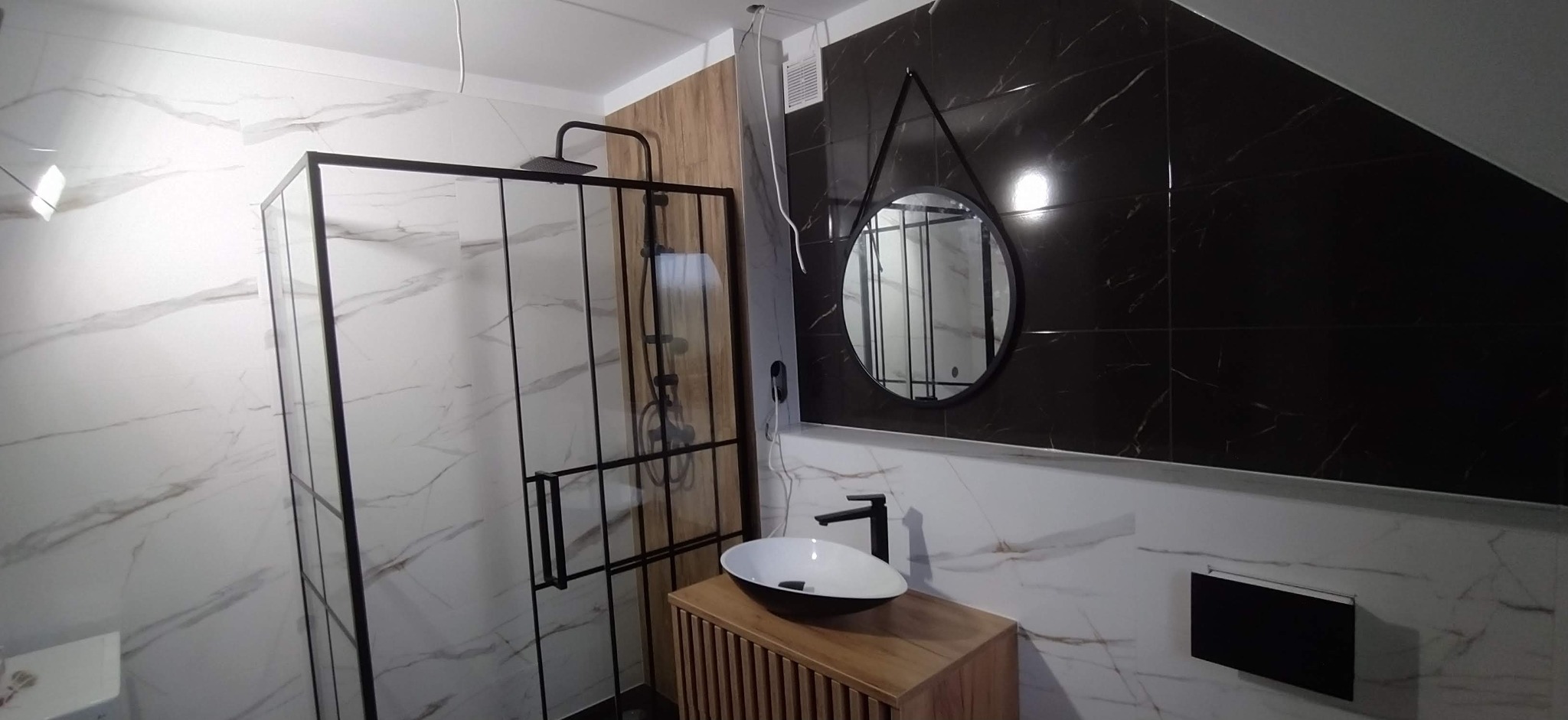 Nowoczesna łazienka z kabiną prysznicową typu walk-in z czarnymi ramami, okrągłym lustrem w czarnej oprawie i umywalką nablatową na drewnianej szafce, wykończona białymi i czarnymi płytkami...