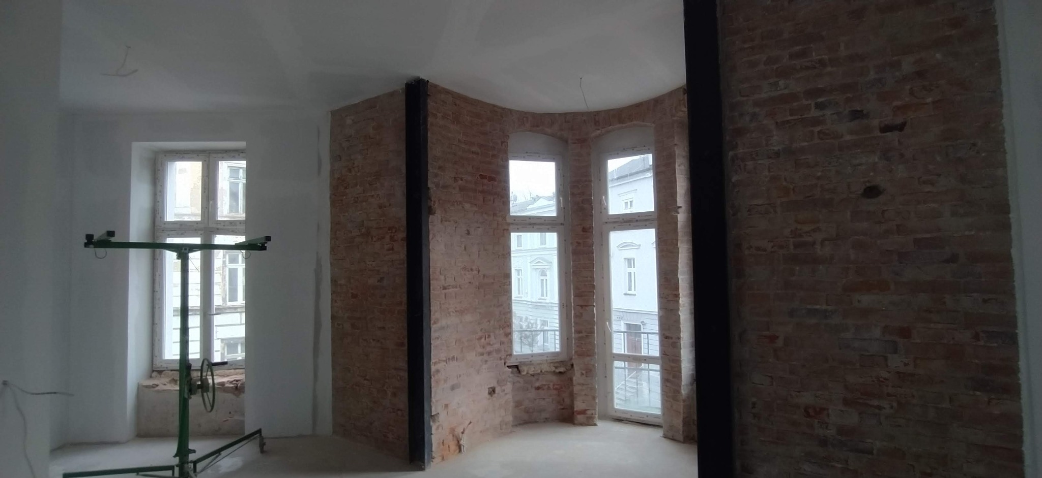 Wnętrze w trakcie remontu z odsłoniętą cegłą, wzmocnieniami konstrukcyjnymi i podnośnikiem do płyt gipsowo-kartonowych przy oknie.