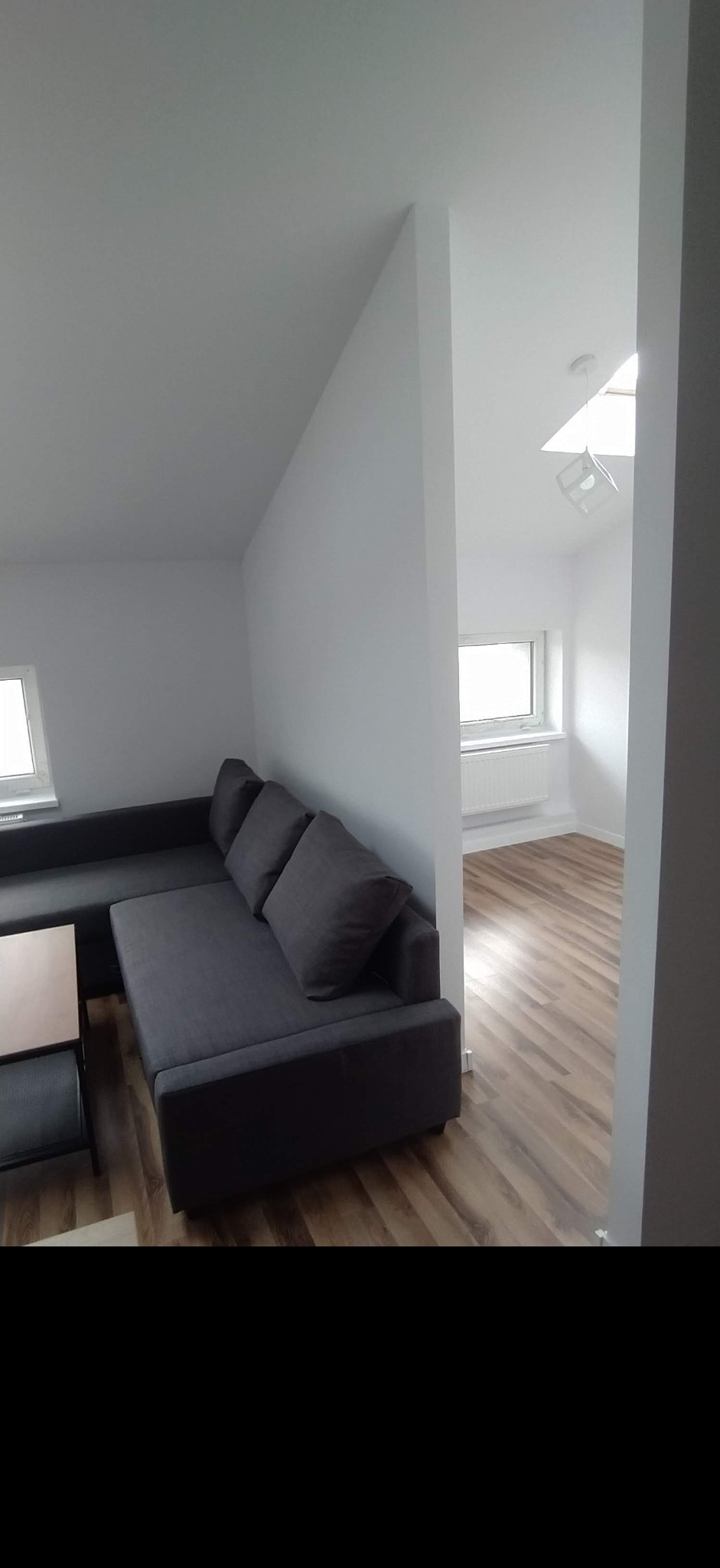 Wnętrze po remoncie: Szary narożnik z poduszkami w salonie z białymi ścianami i drewnianą podłogą, widok na pokój z oknem i grzejnikiem.