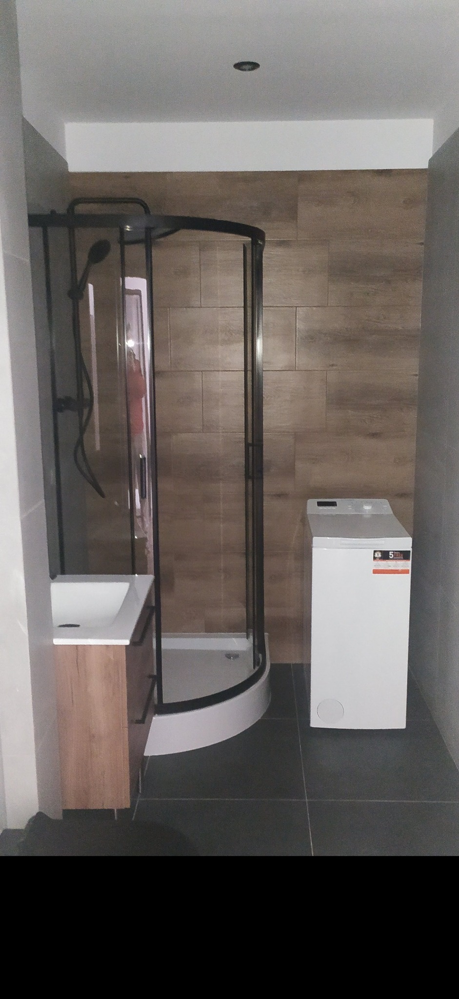 Nowoczesna łazienka z kabiną prysznicową z czarnymi profilami, umywalką z szafką z drewnopodobnym frontem oraz pralką z przodu, wykończona płytkami w odcieniach szarości i brązu.