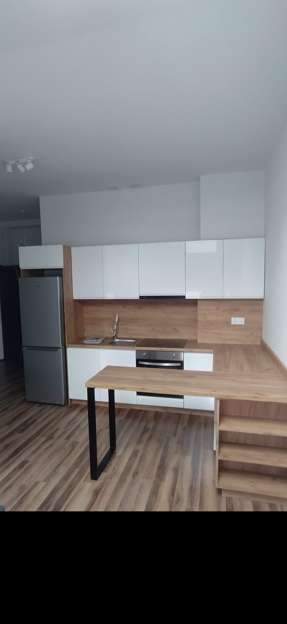 Nowoczesna kuchnia z białymi szafkami, drewnianym blatem i wyspą z czarnymi nogami, podłoga z paneli imitujących drewno.