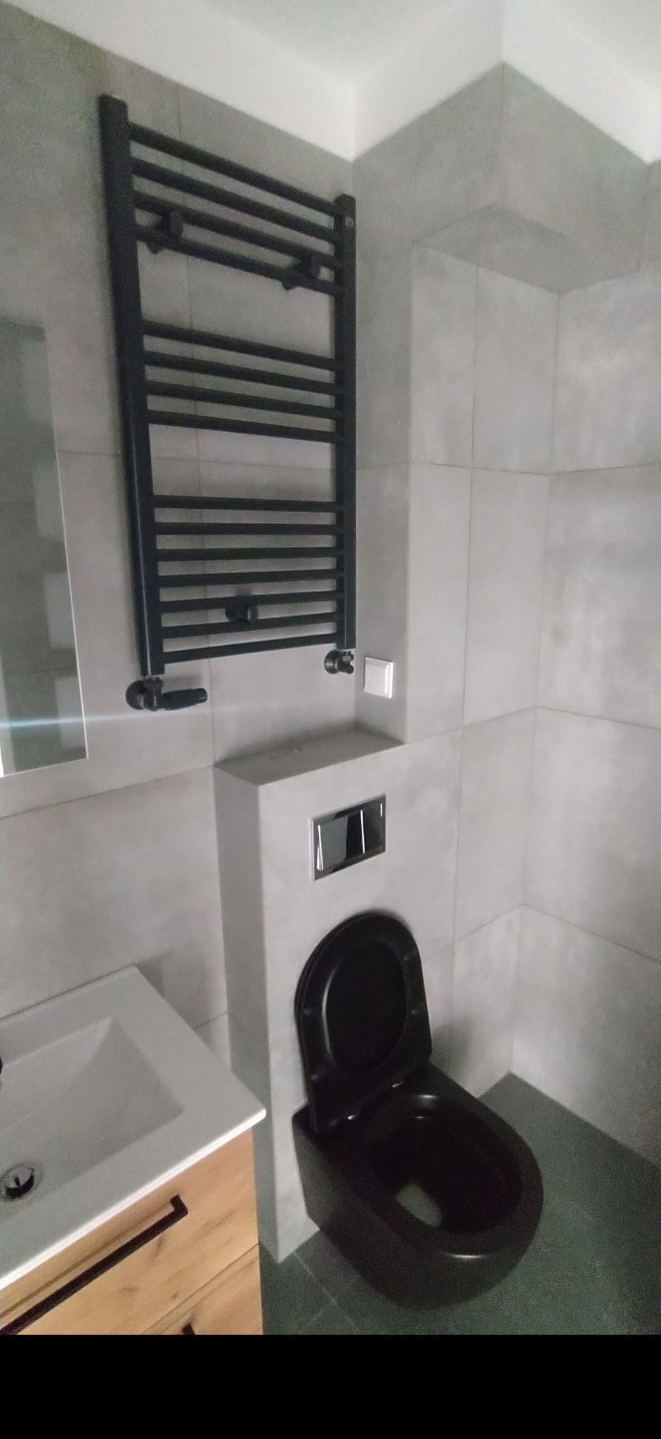 Szara łazienka z wiszącą czarną toaletą, grzejnikiem drabinkowym i szafką pod umywalką z drewnianym frontem.