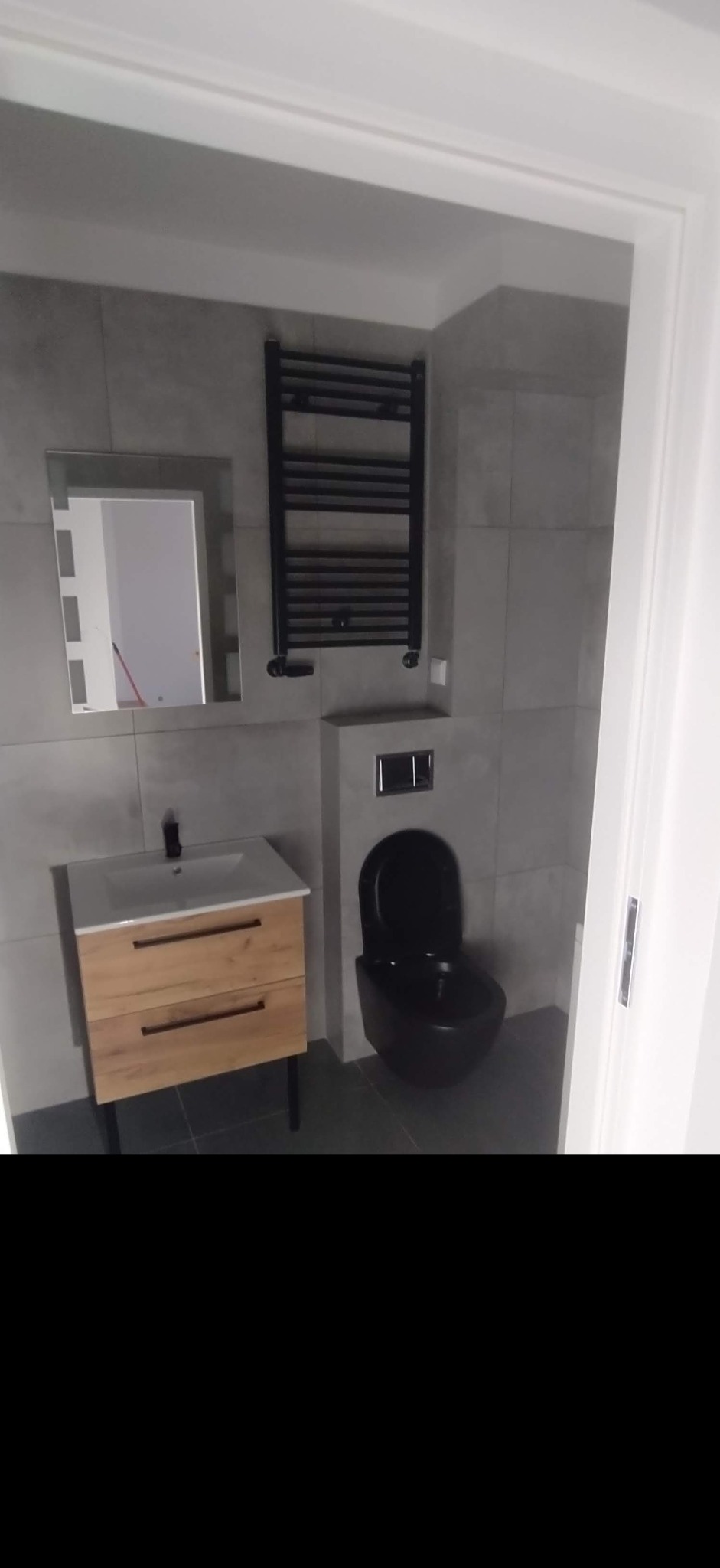 Nowoczesna łazienka z szarymi płytkami, drewnianą szafką pod umywalką, czarną wiszącą toaletą i grzejnikiem drabinkowym, odbicie lustra.