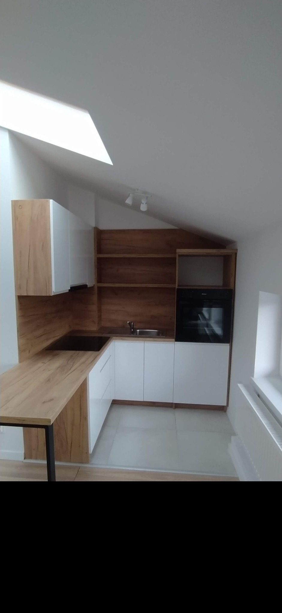 Nowoczesna kuchnia w skosie z białymi frontami i drewnianym blatem oraz otwartymi półkami, oświetlona światłem z okna dachowego.