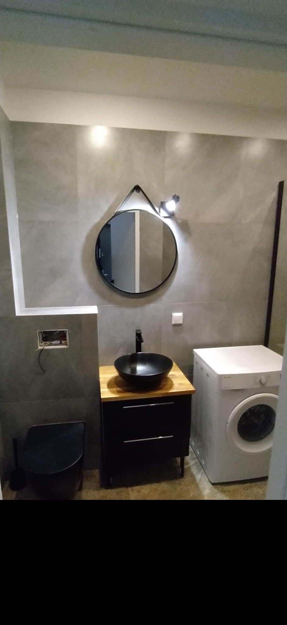 Nowoczesna łazienka z szarymi płytkami, okrągłym lustrem z czarną ramą, czarną umywalką nablatową na drewnianym blacie, czarną toaletą i białą pralką.