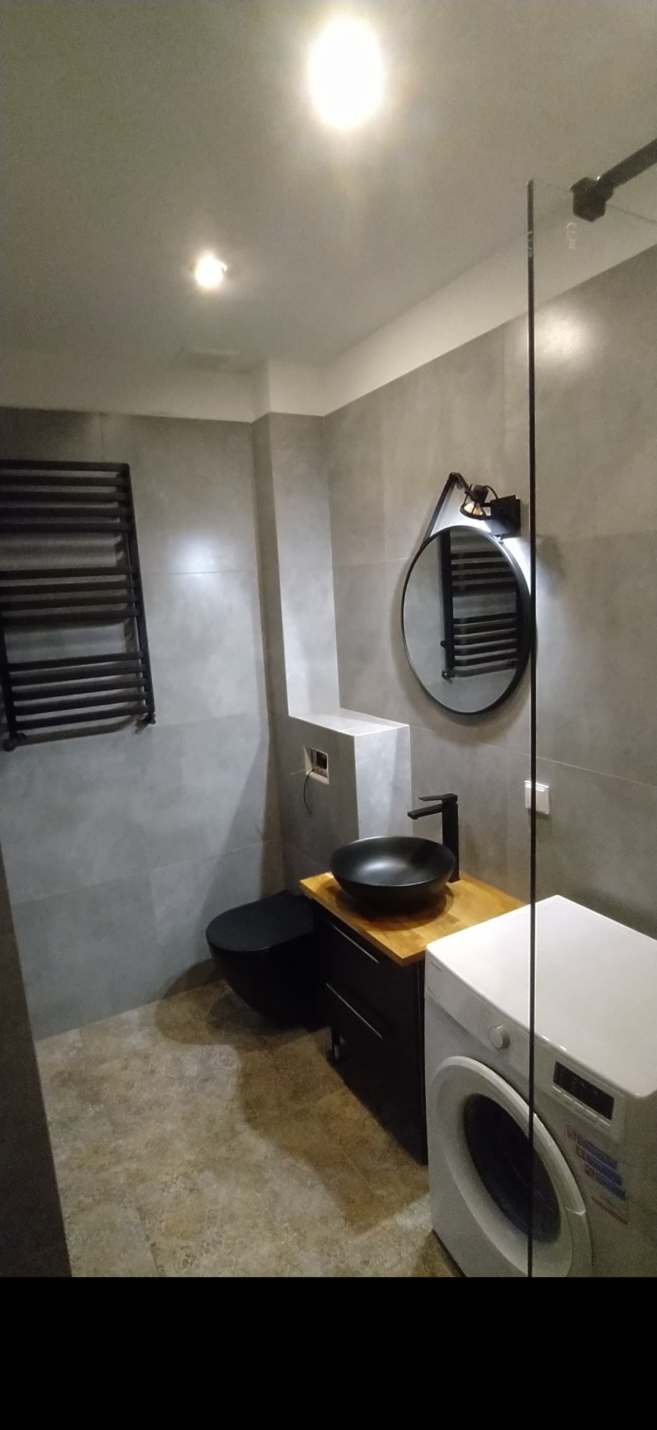 Nowoczesna łazienka w Świętochłowicach z szarymi płytkami, czarną armaturą i okrągłym lustrem z oświetleniem, widoczny grzejnik drabinkowy i pralka.