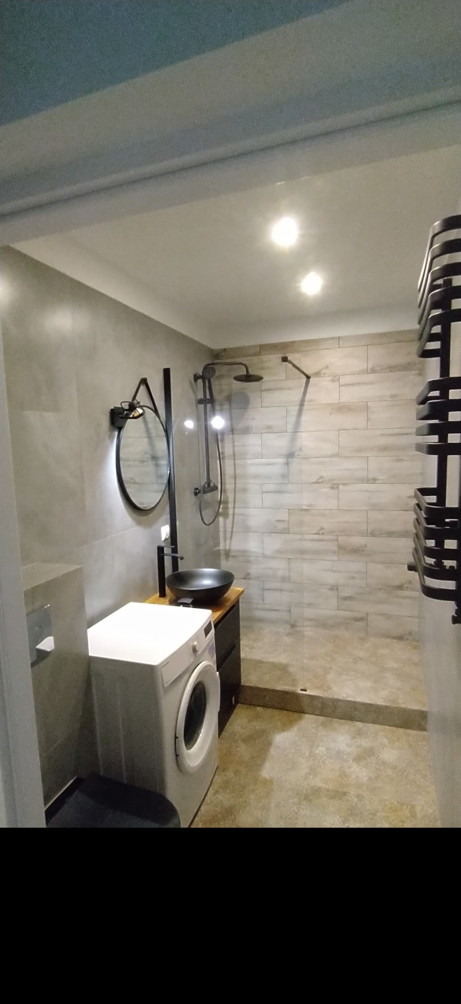 Nowoczesna łazienka z pralką, kabiną prysznicową typu walk-in, umywalką nablatową w kolorze czarnym i okrągłym lustrem z podświetleniem LED.