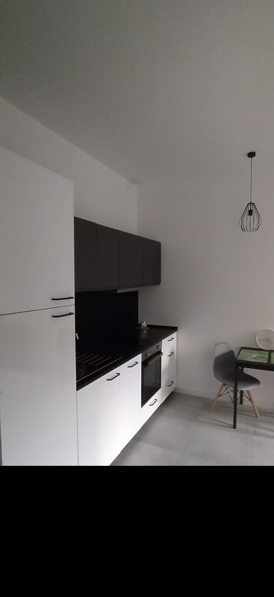 Nowoczesna kuchnia z białymi szafkami dolnymi i ciemnoszarymi górnymi, czarnym blatem i panelem nad blatem, minimalistycznym stołem z szarą krzesłem oraz geometryczną lampą wiszącą.