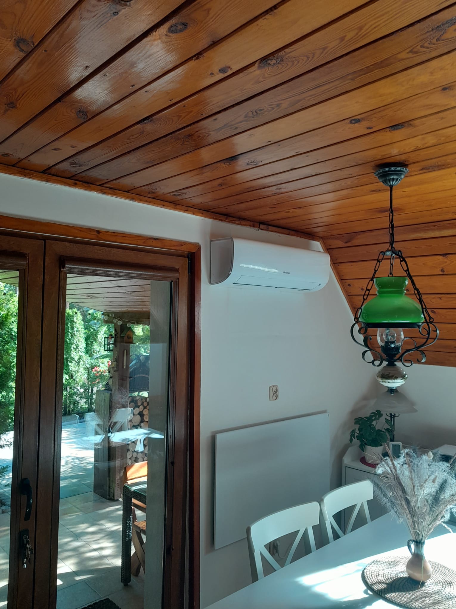 Wnętrze domu z zamontowaną klimatyzacją ścienną, widok na drewniany sufit, przeszklone drzwi balkonowe i ozdobną lampę.