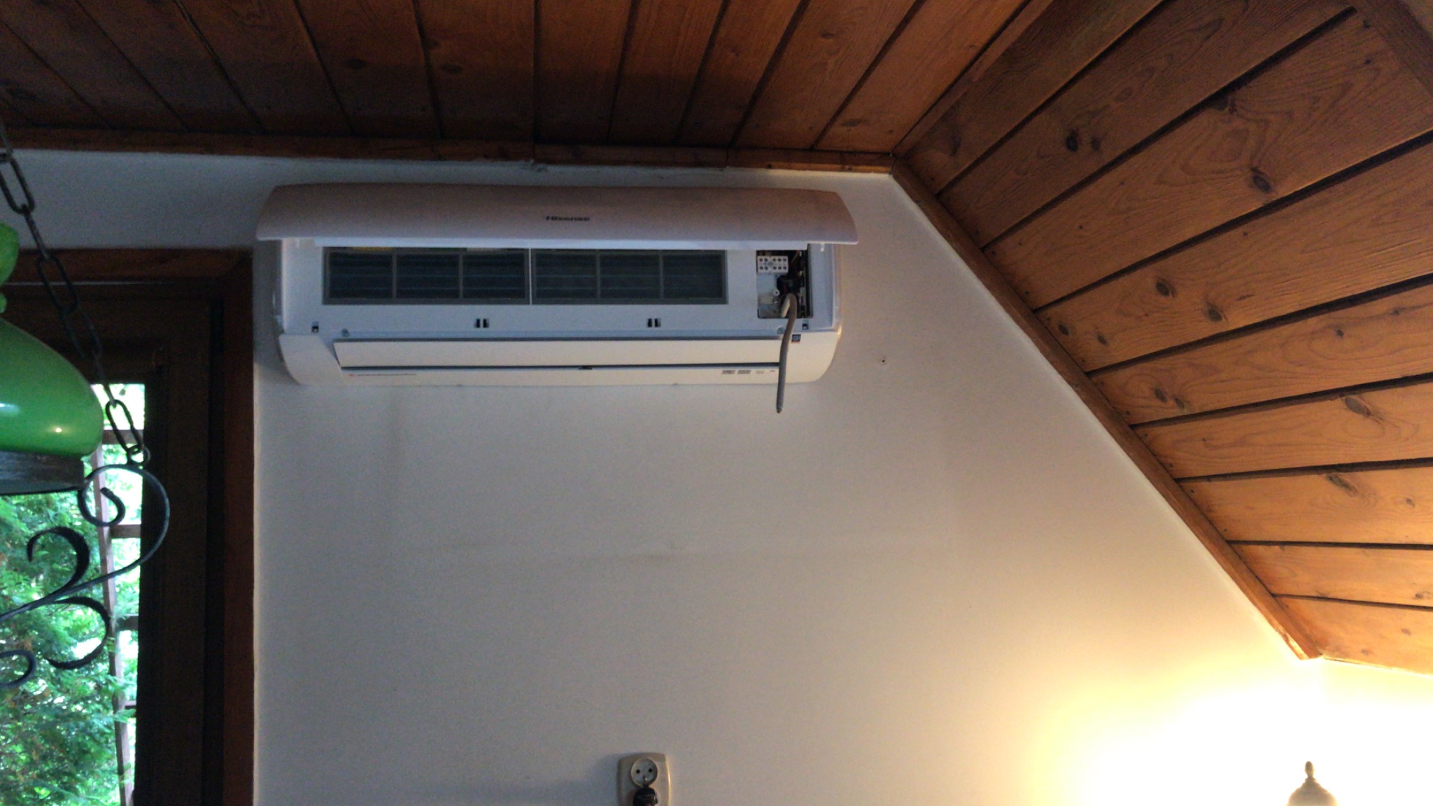 Biała jednostka klimatyzacyjna marki Hisense zamontowana na ścianie w pomieszczeniu z drewnianym sufitem, widoczne otwarte wieko i wystający kabel zasilający.