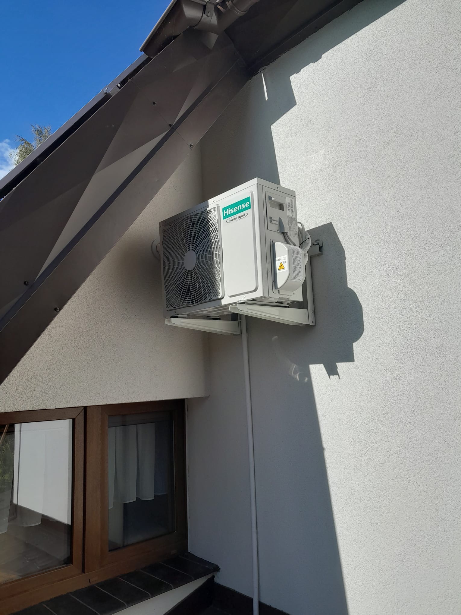 Zewnętrzna jednostka klimatyzacji Hisense zamontowana na białej ścianie domu pod okapem dachu, widoczne okno z brązową ramą.