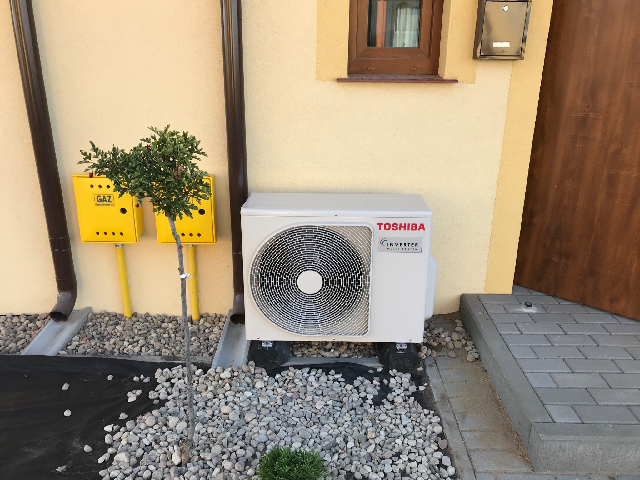 Zewnętrzna jednostka klimatyzacji Toshiba Inverter Multi System zamontowana na zewnątrz domu, na podłożu z kamieni, obok skrzynki gazowej i rury spustowej.