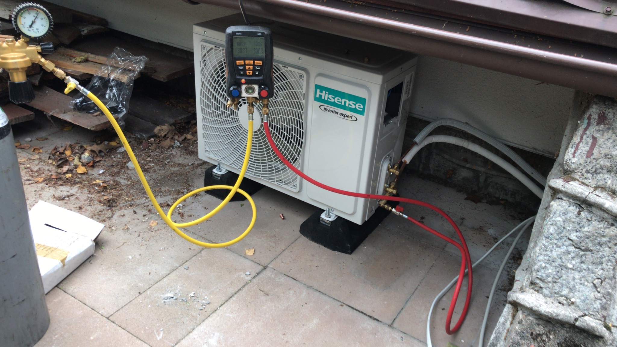 Podłączenie manometru cyfrowego do jednostki zewnętrznej klimatyzacji Hisense Inverter Expert na szarym chodniku, widoczne przewody i butla z gazem chłodniczym.