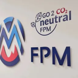 Trójwymiarowe logo firmy z elementami w kolorach niebieskim i czerwonym, obok napis 'GO 2 CO2 neutral FPM' i 'FPM' na białej ścianie.