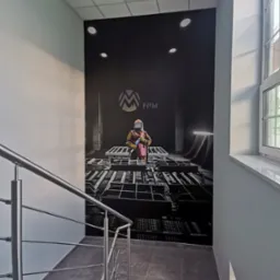 Klatka schodowa z fototapetą przedstawiającą spawacza w kombinezonie ochronnym i logo FPM na czarnym tle, widok z góry na schody.