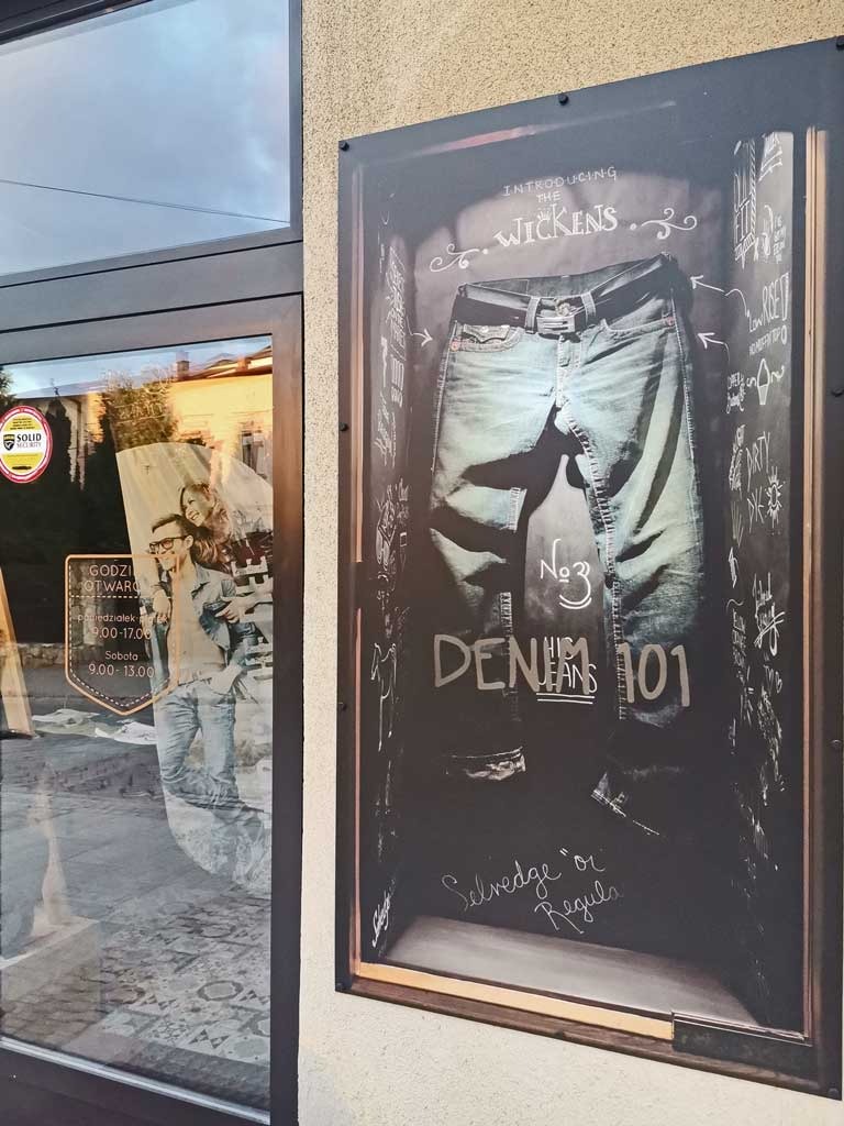 Zewnętrzny plakat reklamowy jeansów marki Wickens z efektem tablicy kredowej, umieszczony obok przeszklonych drzwi wejściowych sklepu.