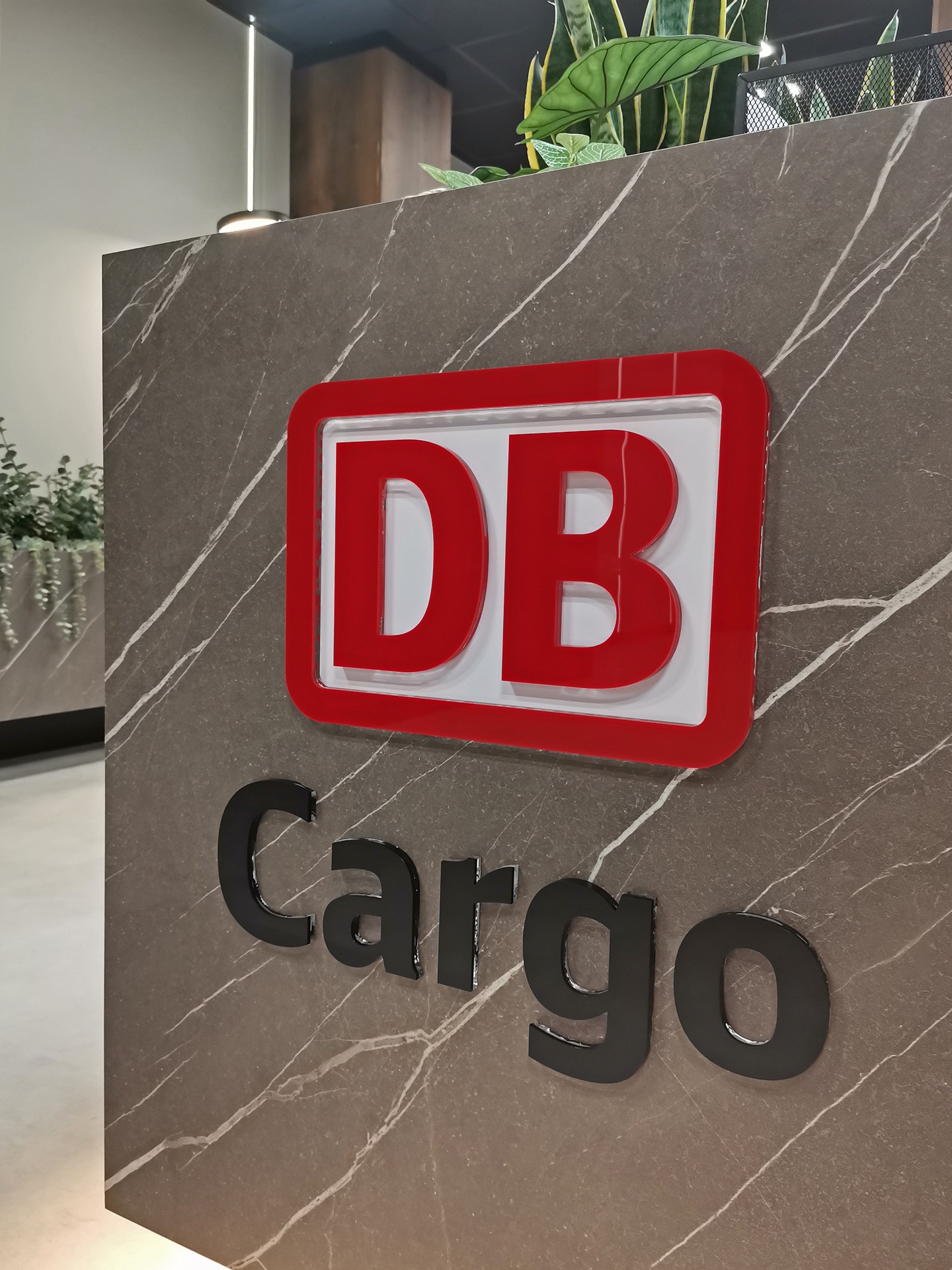 Logo DB Cargo z czerwonym obrysem na marmurowej ścianie w biurze, widoczne rośliny w tle.