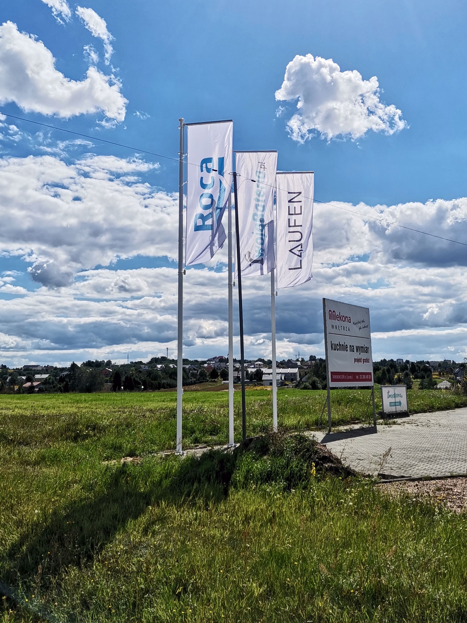 Flagi reklamowe Roca i Laufen na tle zielonego pola i błękitnego nieba z chmurami, obok tablica reklamowa firmy Mlekona Wnętrza z ofertą kuchni na wymiar.