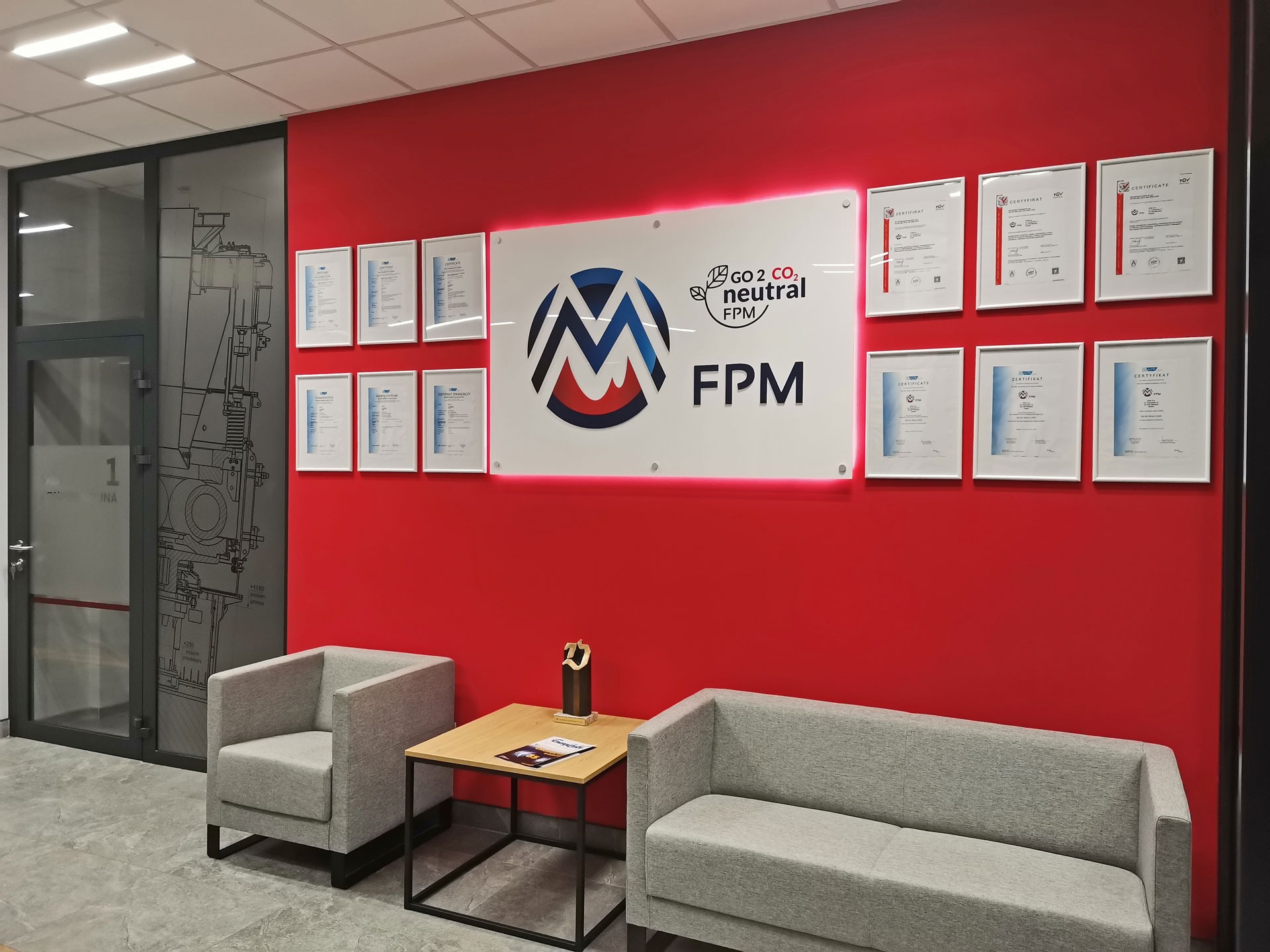 Wnętrze biura z czerwoną ścianą, na której wiszą oprawione certyfikaty i logo firmy FPM z hasłem 'GO 2 CO2 neutral'. Dwa szare fotele i sofa oraz stolik kawowy z nagrodą i katalogiem tworzą strefę...