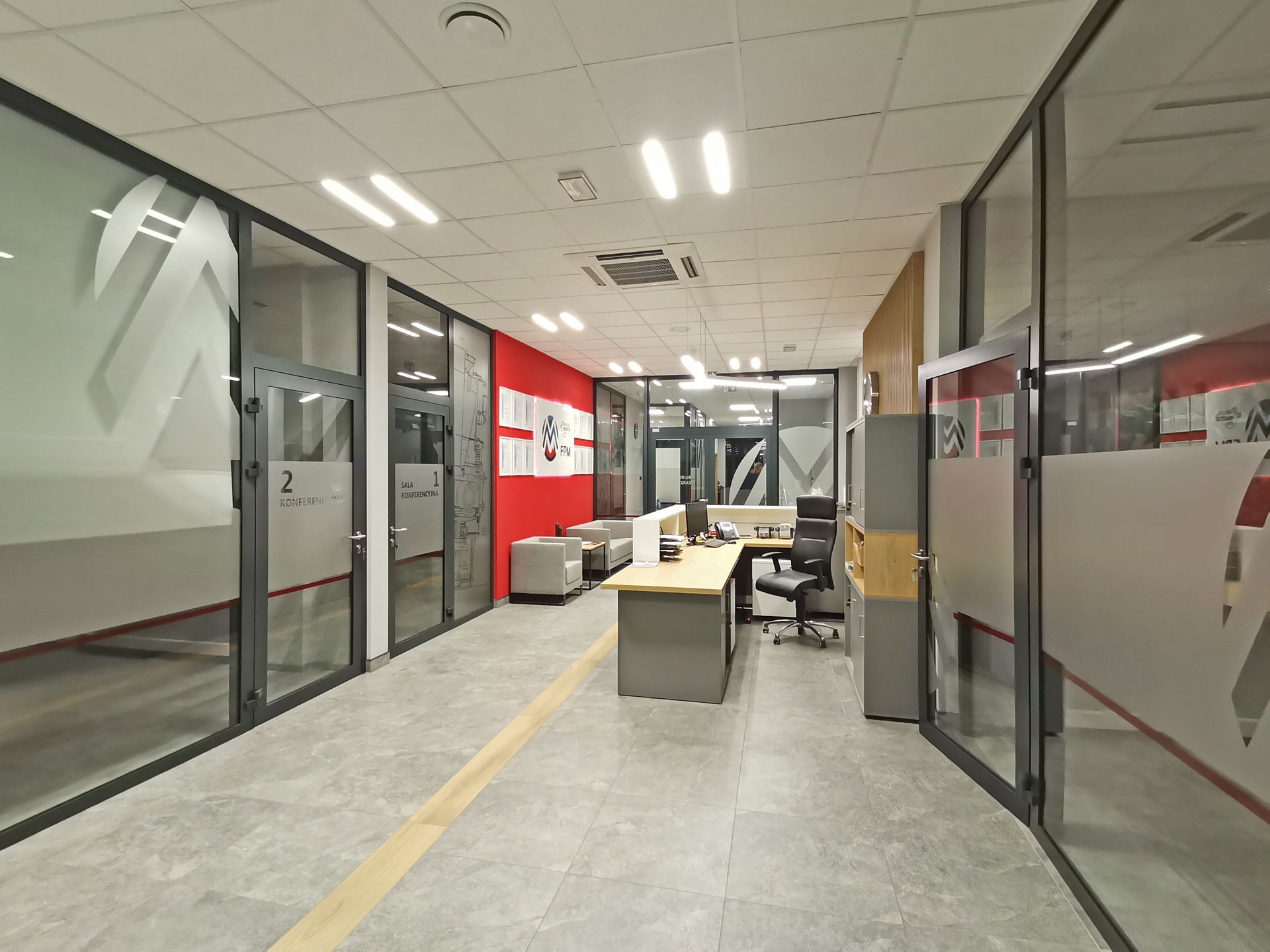 Wnętrze biura z przeszklonymi ścianami, na których umieszczono logo firmy wykonane metodą piaskowania, widoczne biurko i krzesło biurowe oraz strefa relaksu z sofami.