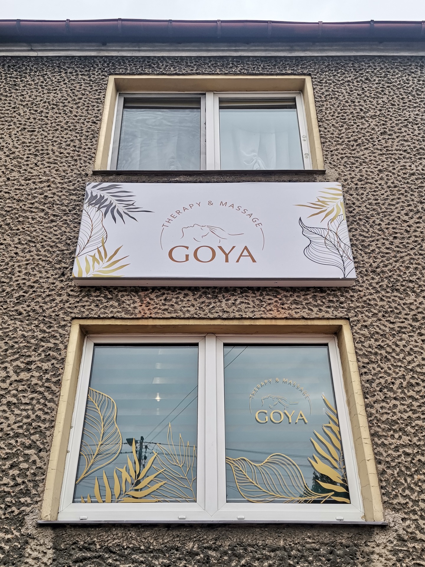 Elewacja budynku z szyldem 'Goya Therapy & Massage' oraz dekoracyjnymi naklejkami z motywem liści na oknach, widok z dołu.