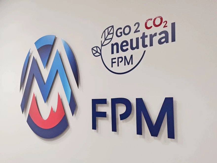 Trójwymiarowe logo firmy z elementami w kolorach niebieskim i czerwonym, obok napis 'GO 2 CO2 neutral FPM' i 'FPM' na białej ścianie.