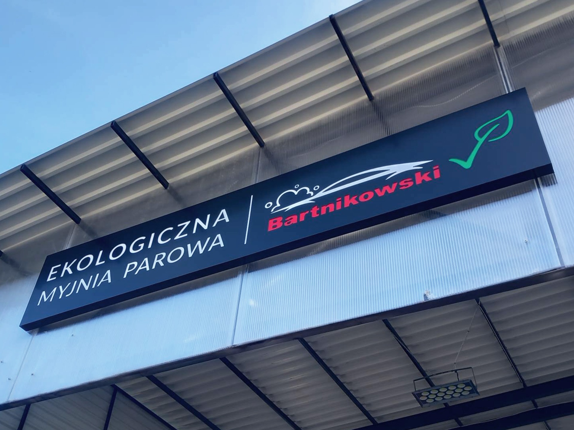 Czarny szyld reklamowy z napisem 'Ekologiczna Myjnia Parowa Bartnikowski' z logo firmy i zielonym symbolem liścia, zamontowany na jasnej elewacji budynku.