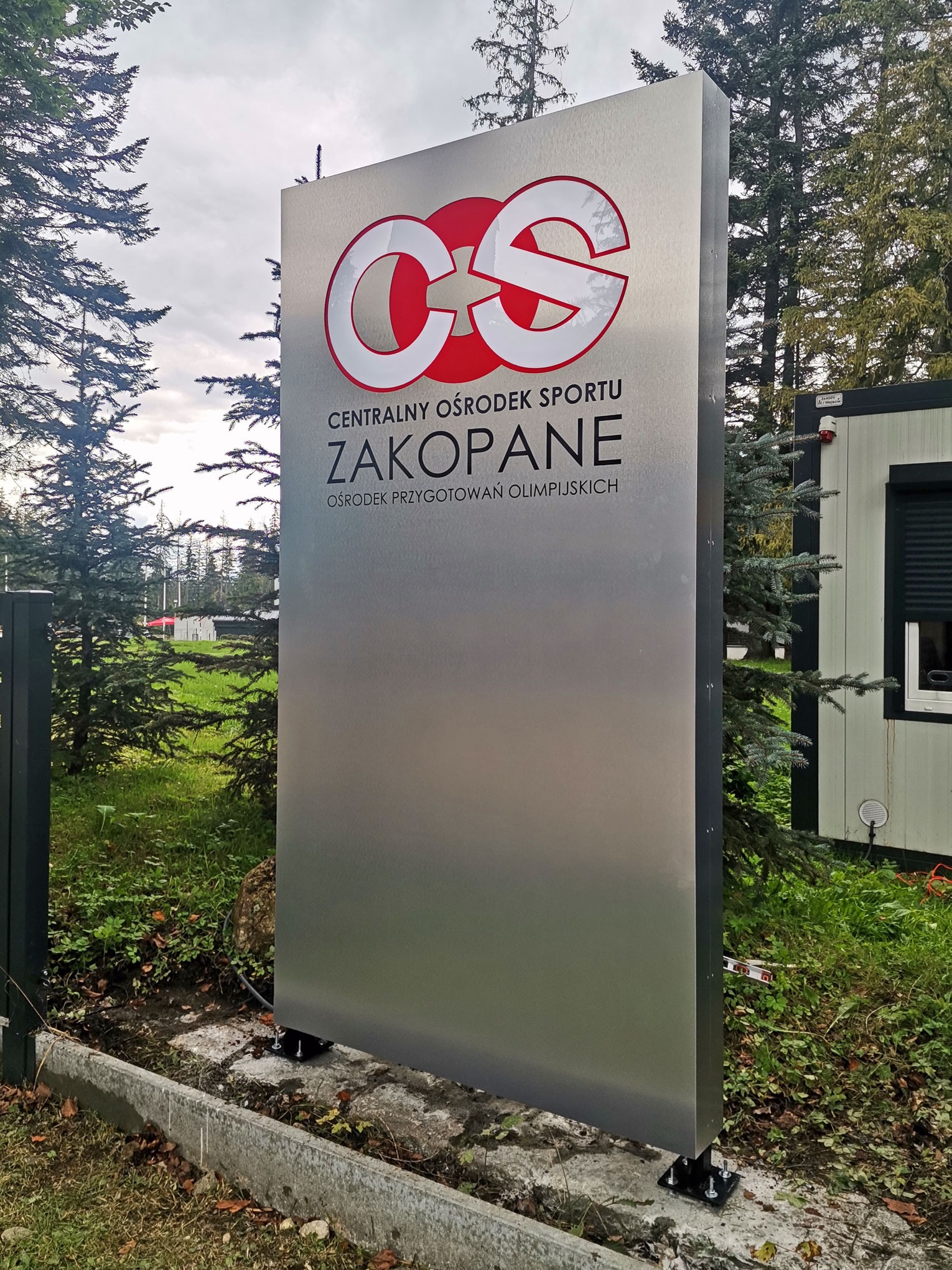Metalowy pylon informacyjny z logo i nazwą Centralnego Ośrodka Sportu w Zakopanem, umieszczony na zewnątrz w otoczeniu zieleni i drzew.