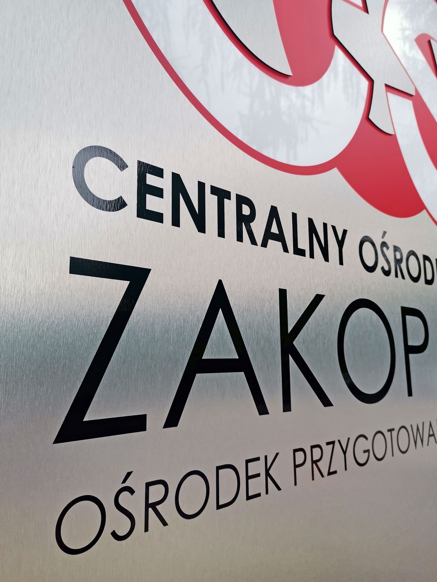 Metalowa tablica informacyjna z napisem 'Centralny Ośrodek Zakopane Ośrodek Przygotowań' w kolorach czarnym, czerwonym i białym, widoczna z bliska pod kątem.