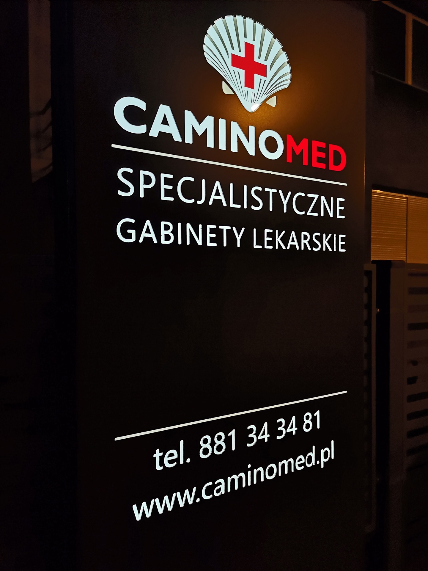 Czarny, podświetlany szyld z nazwą 'CAMINOMED', logotypem muszli z czerwonym krzyżem, napisem 'Specjalistyczne Gabinety Lekarskie', numerem telefonu i adresem strony internetowej.