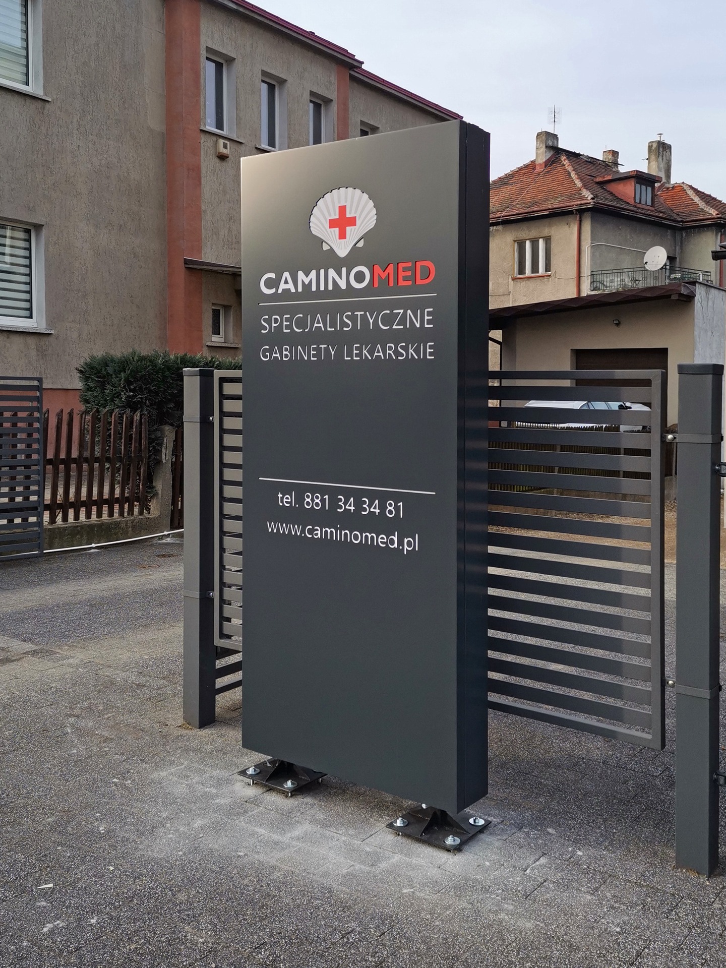 Pionowy szyld reklamowy gabinetu lekarskiego CaminoMed z logo muszli z czerwonym krzyżem, umieszczony na zewnątrz budynku, widoczny zza metalowej bramy.