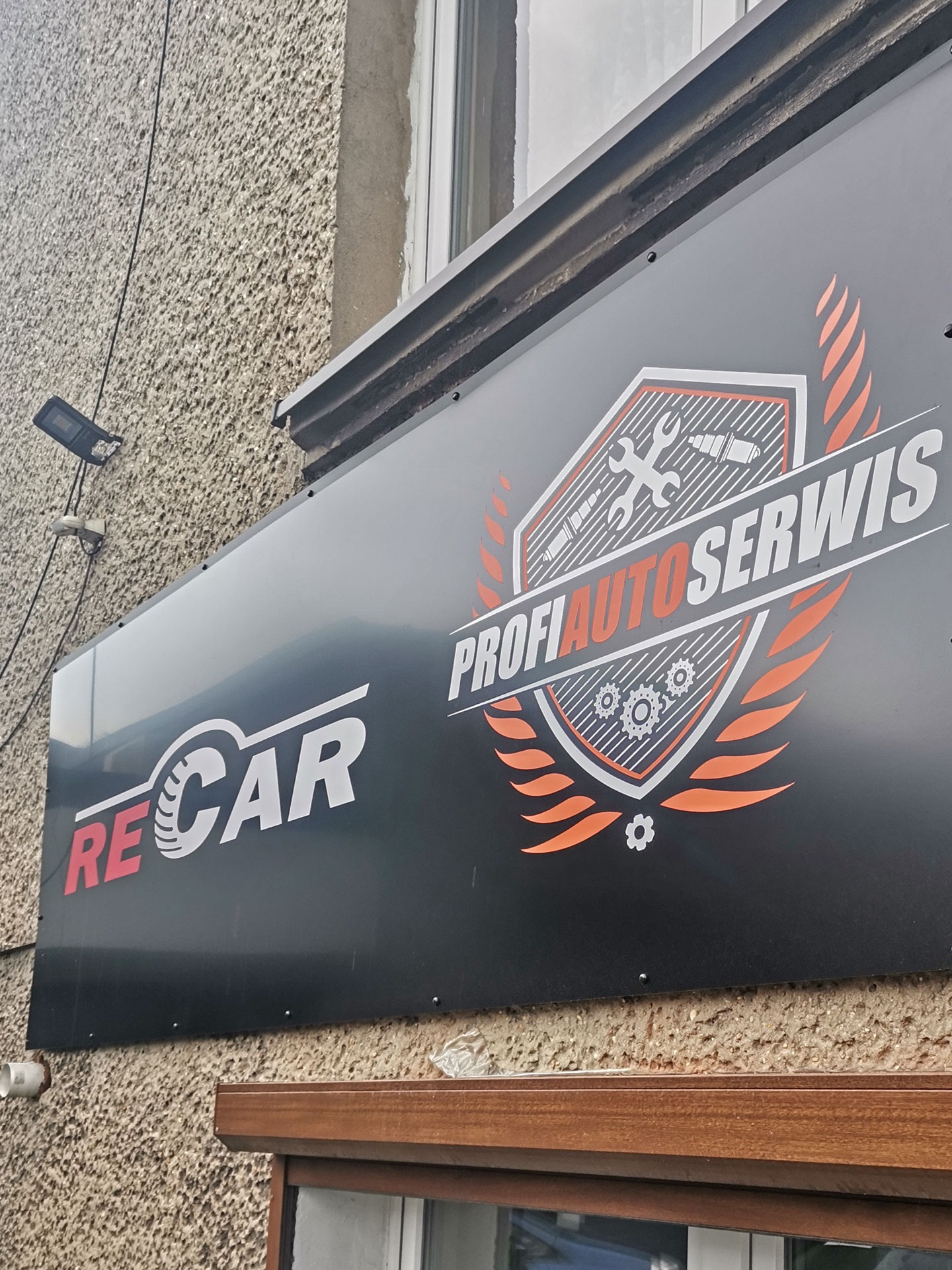 Szyld reklamowy z logo 'RECAR' oraz 'PROFI AUTOSERWIS' z grafiką narzędzi warsztatowych i płomieni, zamontowany na elewacji budynku.