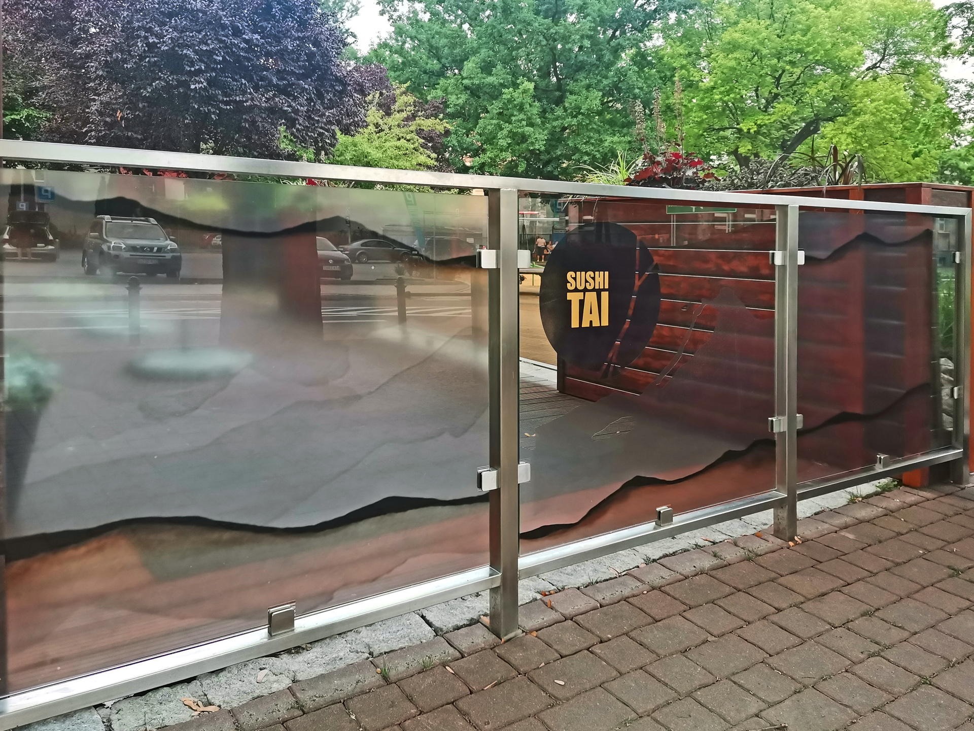 Szklana balustrada z motywem górskim i logo 'Sushi Tai' na drewnianym tle, widok z zewnątrz na ulicę z przejeżdżającymi samochodami.