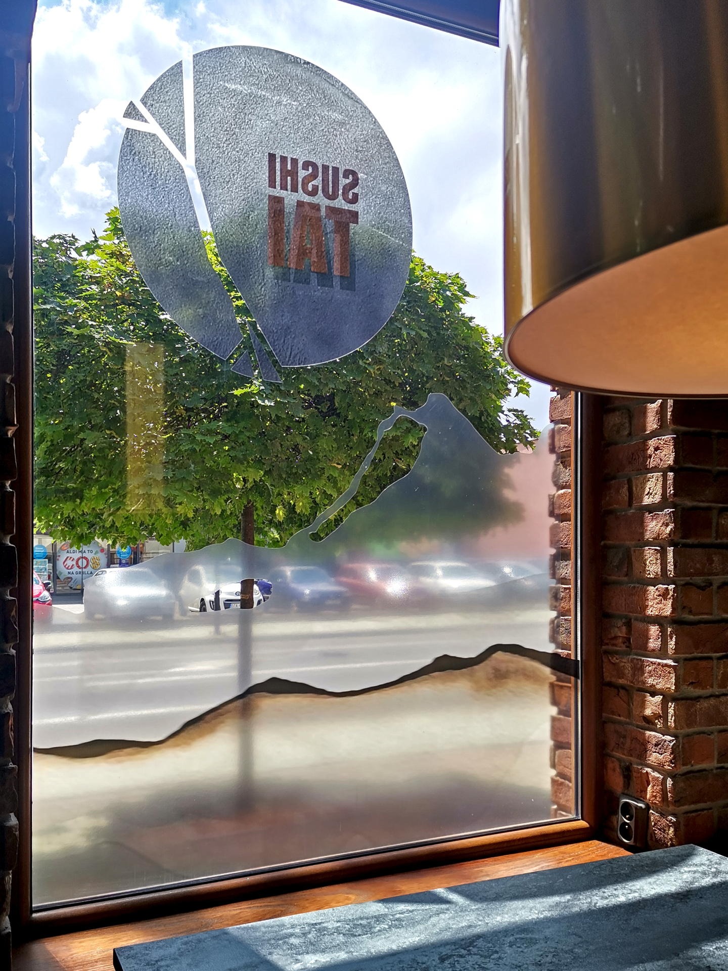 Ozdobna folia na oknie restauracji sushi z motywem górskim i logo, widok na ulicę z drzewami i samochodami, ceglana ściana w tle.