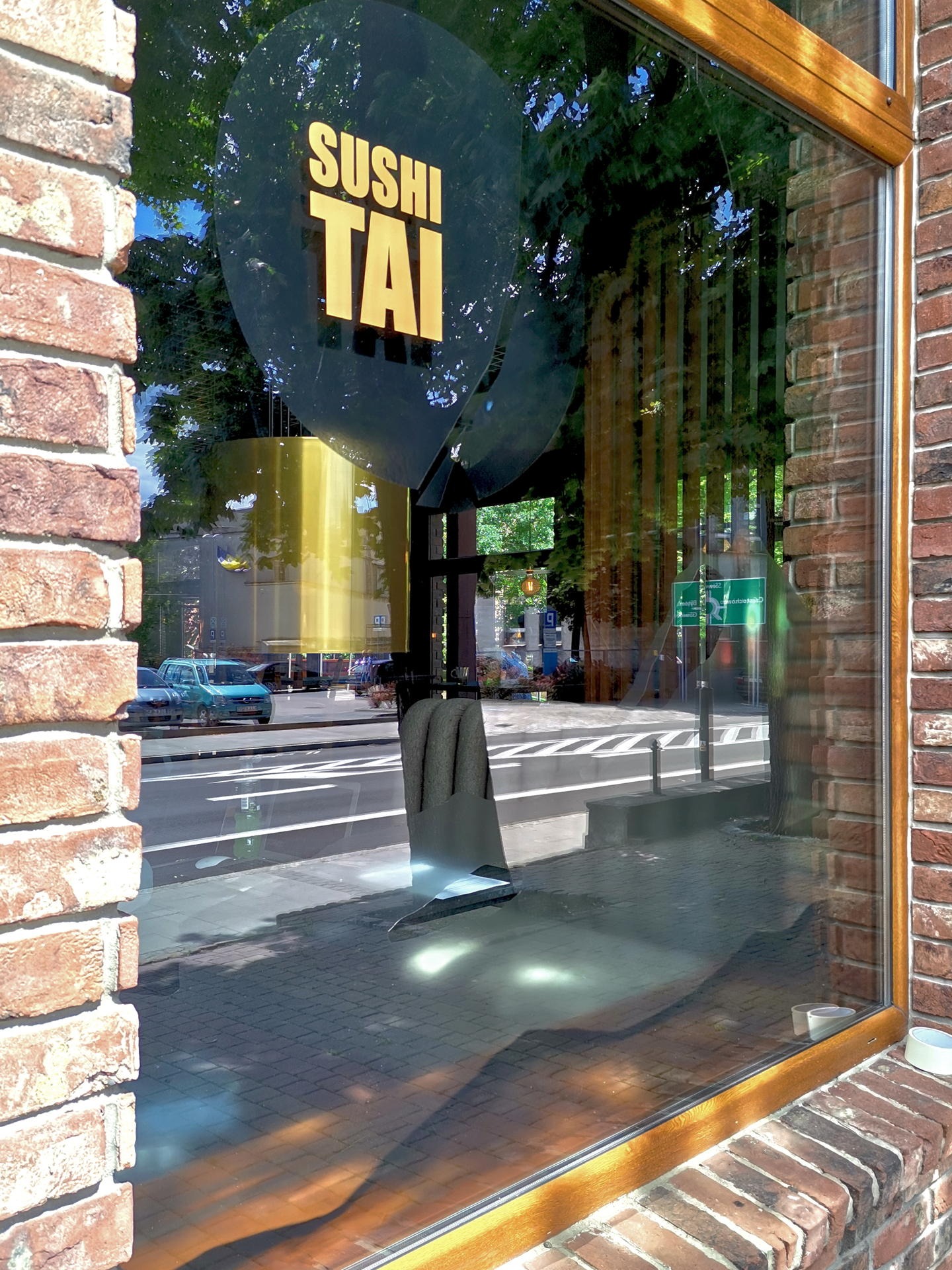 Witryna restauracji Sushi Tai z logo na tle ulicy i drzew, odbicie chodnika i krzesła w szybie, ceglana ściana z drewnianą ramą okienną.