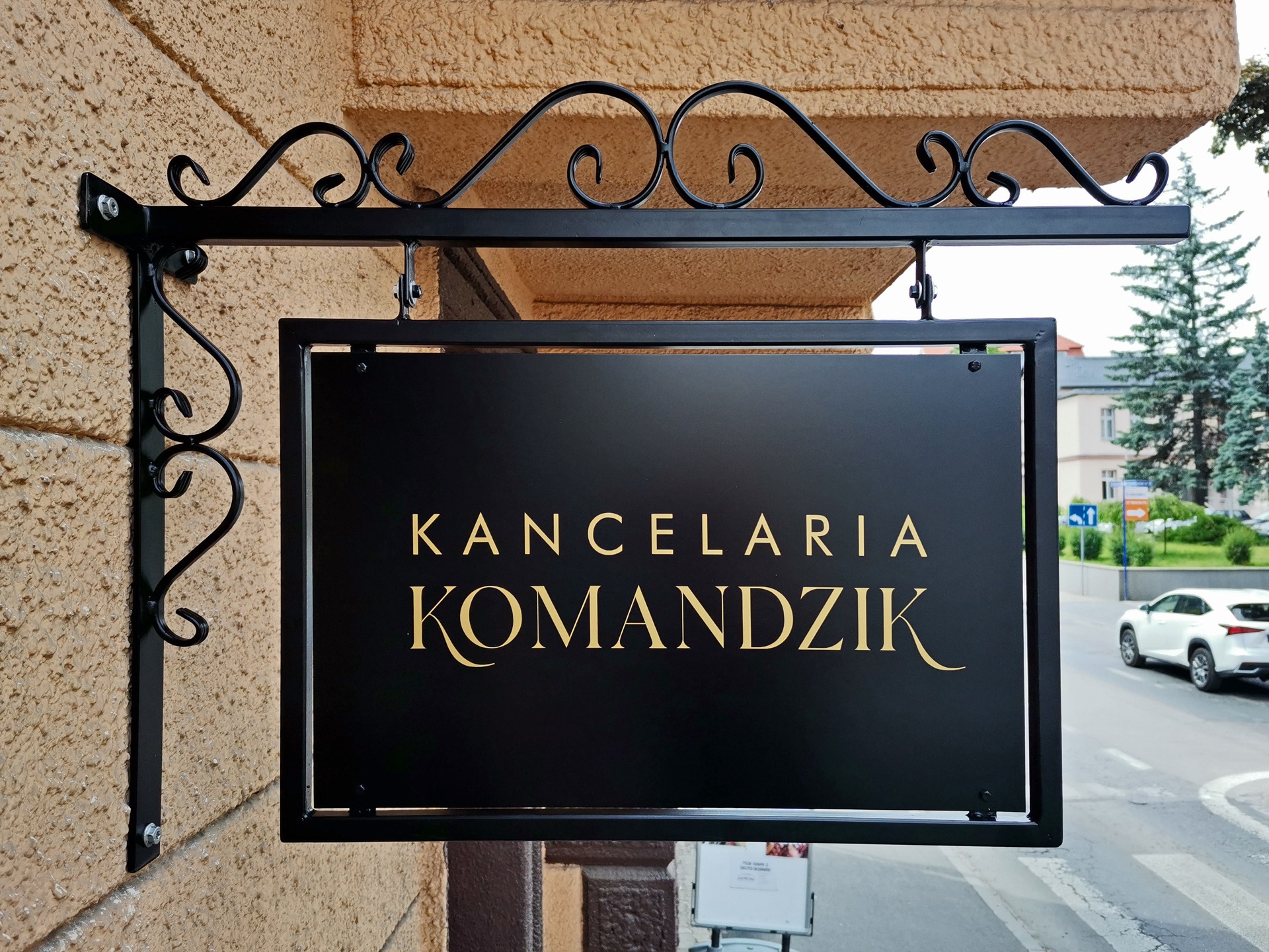 Elegancki szyld z kutego żelaza z napisem 'Kancelaria Komandzik' w kolorze złotym, zamontowany na elewacji budynku.
